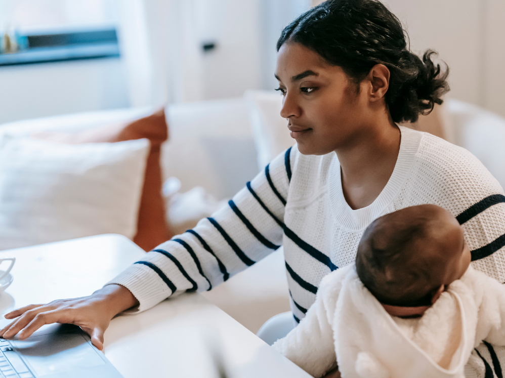 A stock image showing a mother working from home on a laptop, while holding her baby.