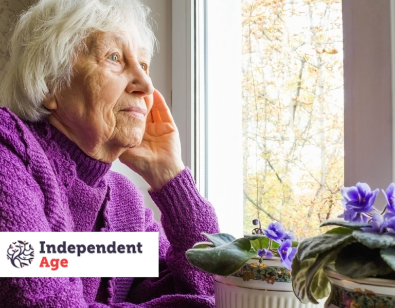 A stock image showing an older woman with white hair looking out of her window. She's wearing a purple cardigan, and she looks thoughtful. The Independent Age logo is there too.