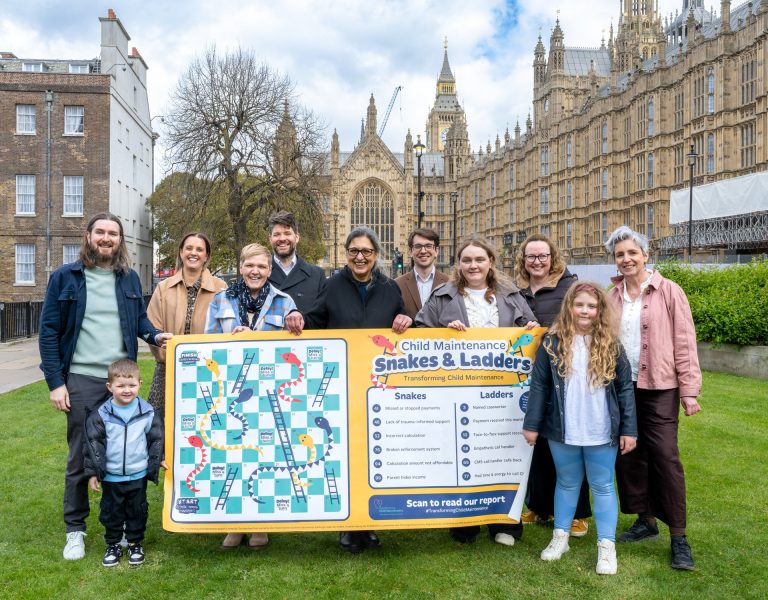 A group of men and women from the Transforming Child Maintenance partnership on St Stephen's Green outside the Palace of Wesminster. They're holding up a snakes and ladders game to represent that they used to show MPs how difficult the child maintenance system is to navigate.
